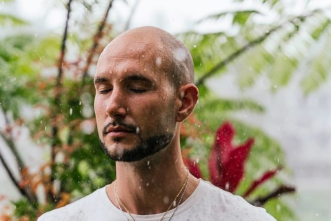 A man stands with closed eyes, embracing the gentle rainfall. Surrounded by lush greenery, he