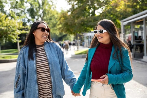A joyful Latin lesbian couple strolls hand in hand through a lush, sunlit park. Both are wearing