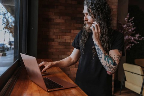 A bearded man with long hair and tattoos uses a laptop while talking on the phone, positioned near