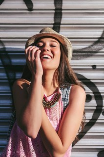A cheerful woman in a hat and pink top enjoys a sunny day, standing against a metal shutter