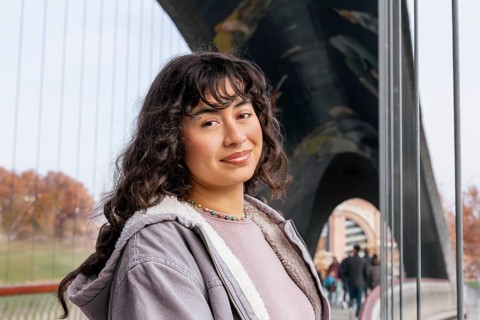 A young Latin woman with curly hair and a relaxed smile, casually dressed, stands confidently on a