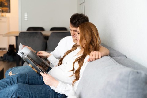 A young couple sits together on a cozy sofa, embracing and reading a magazine. The expectant mother