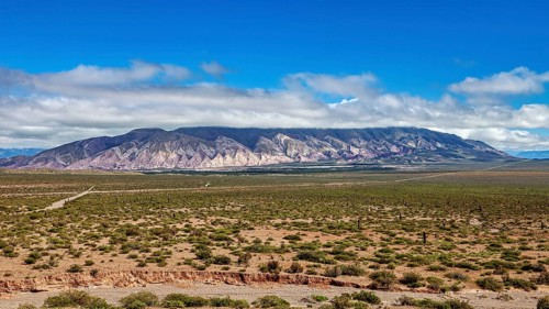 Wide plain in front of a mountain range under a blue, cloudy sky, the landscape and colorful rock