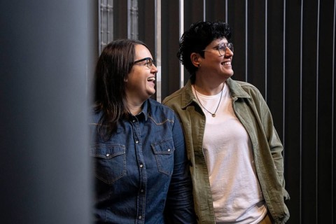 A lesbian couple enjoys a moment of laughter together. Both wear casual attire and glasses,
