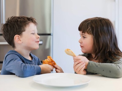 Two children joyfully share a meal at a kitchen table, holding slices of pizza. The scene captures