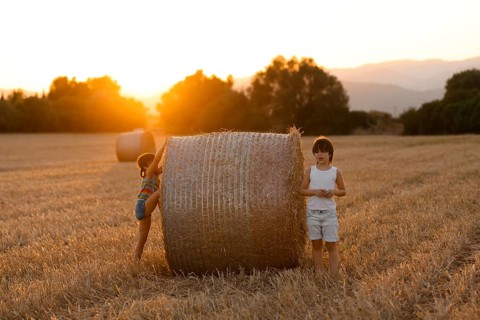 A young boy and girl play near a large hay bale in a field bathed in the warm light of a setting