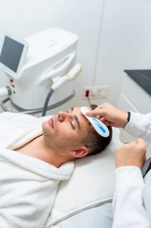 Male client relaxing wrapped in a white robe on a treatment bed while a professional cosmetologist