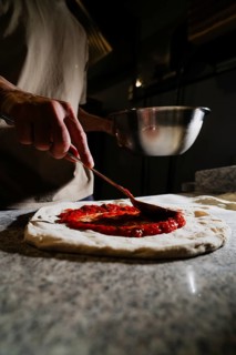 A person spreading fresh tomato sauce on a pizza dough in a dimly lit kitchen. The focus is on the