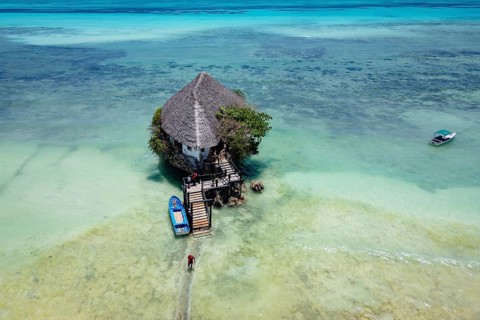 Aerial view of the building surrounded by turquoise water and white sand beach with boats under a