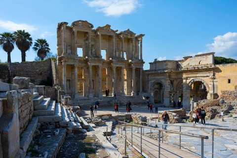 Ancient library façade with palm trees and tourists surrounded by historical ruins, Celsus Library,