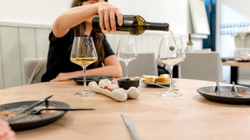 A woman pours white wine into a glass at a restaurant dinner setting. The table is elegantly set