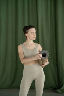 Woman in beige fitness attire holding a rolled yoga mat, set against a green curtain backdrop.