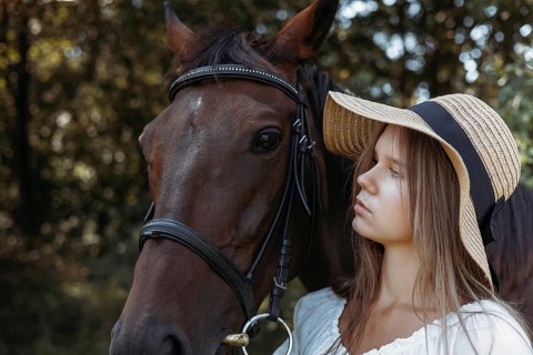 A young girl with long hair and a wide brimmed straw hat stands closely next to a brown horse