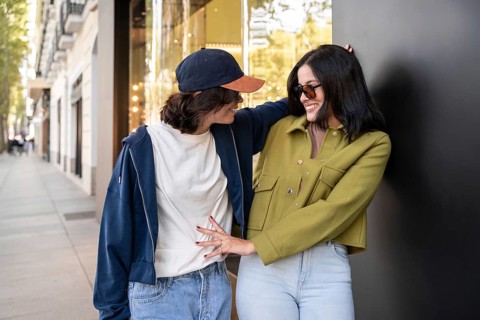 A happy lesbian couple embraces while walking down a sunny city street. They appear relaxed and