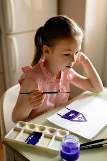A young girl in a pink dress concentrates intently while painting a simple house with purple