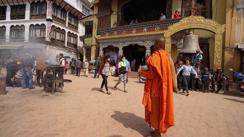 A monk dressed in orange walks past a lively place of prayer with traditional architecture, Bodnath