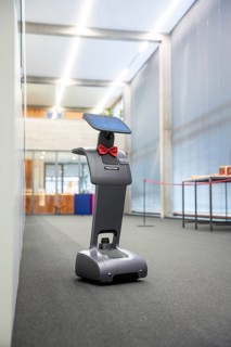 A robot-like device with a red ribbon stands on a carpeted floor in an illuminated office,