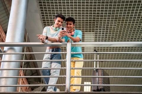 A joyful gay couple stands at a railing, sharing a moment and looking at a smartphone. They appear