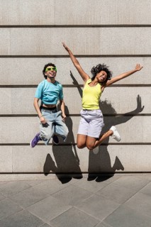 A multiethnic LGBTQ+ couple joyfully jumps in bright, colorful outfits against an urban backdrop,