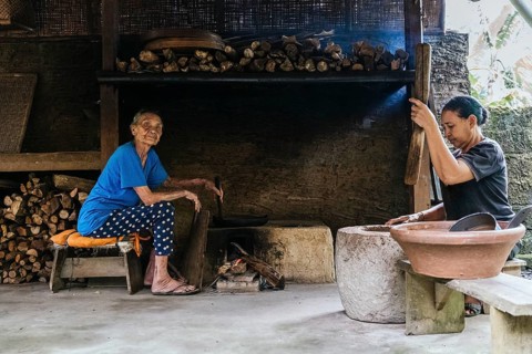 Two women work in a rustic kitchen, surrounded by firewood and traditional cookware. One prepares