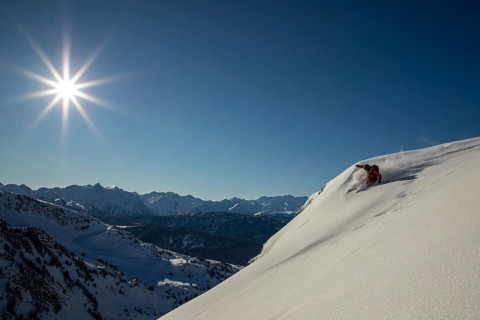 A skier glides down a steep, pristine snow-covered mountain slope under a clear blue sky The sun