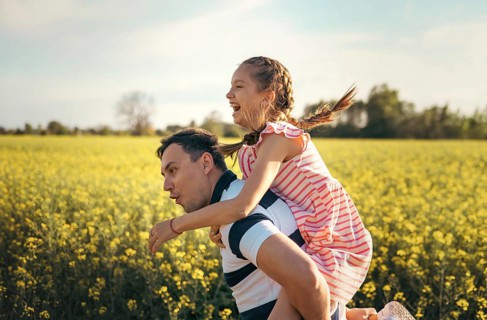 A joyful father carries his daughter on his back as they laugh together in a sunlit flower field.