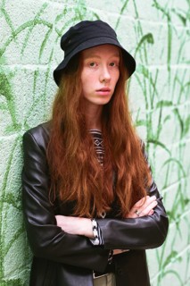 Young woman with long red hair in a stylish black hat and leather jacket in a vibrant green