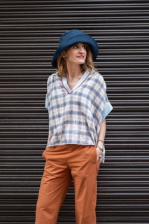A stylish woman in a dark background, wearing a blue hat, plaid shirt, and orange pants, exuding