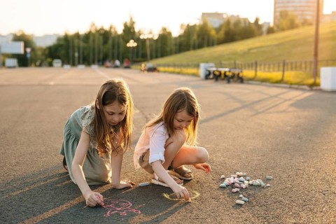 Two young girls joyfully create chalk art on a warm, sunny day in the park. Sitting on the pavement