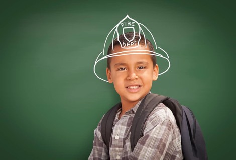 Young Hispanic Student Boy Wearing Backpack Front Of Blackboard with Fireman Helmet Drawn In Chalk