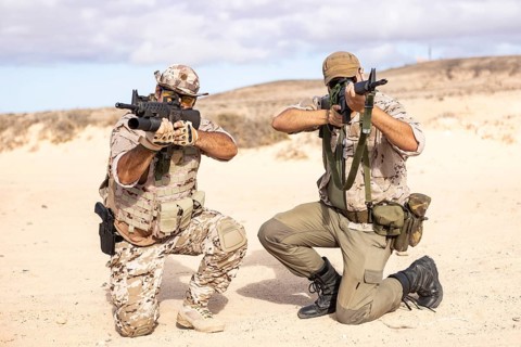 Two military soldiers kneel in a desert landscape, aiming rifles. They wear tactical gear and