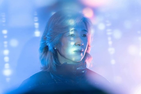An elderly Asian woman interacts with a colorful metaverse interface, surrounded by ethereal blue