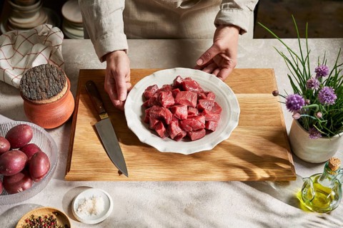 A butcher presents a plate of fresh, red meat cubes on a rustic wooden board, surrounded by kitchen