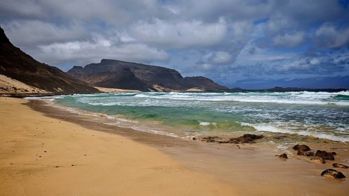 Remote, secluded sandy beach by the sea with waves and mountains in the background under cloudy