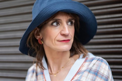 A fashion woman wearing a blue hat and plaid shirt stands against a dark background, exuding a