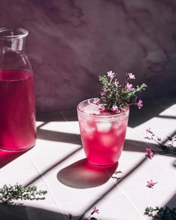 A glass of vibrant pink lemonade, garnished with delicate purple flowers, sits beside a transparent