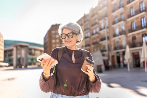 Senior woman walking in a sunny city square, smiling while listening to music with headphones,
