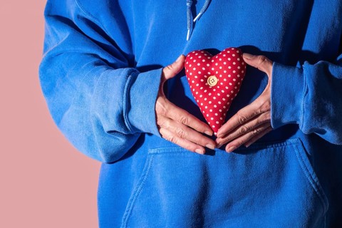 Close up of a person in a blue hoodie holding a red heart shaped pillow with white polka dots and a
