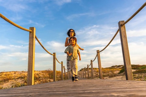 Mother and son joyfully running on a wooden boardwalk leading to sand dunes, enjoying a summer