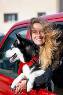 A smiling woman with her husky dog leans out of a red car in excitement. The dog's tongue is out,