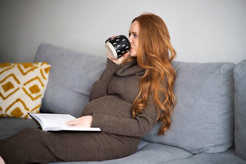Pregnant woman relaxing on a cozy gray sofa, enjoying a warm cup of coffee while reading a book.
