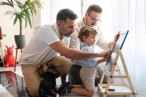 A gay couple lovingly teaches their young son to draw at home, engaging him in creative activities.