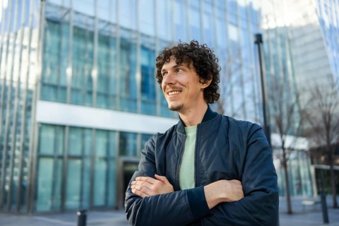 A cheerful man stands confidently with crossed arms, smiling in an urban setting. The background