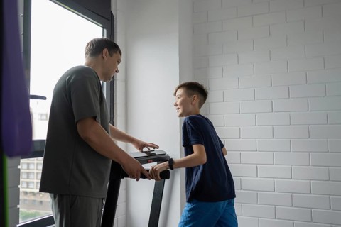 A father and son share a joyful moment exercising on a treadmill at home. Natural light streams