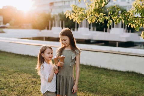 Two young girls enjoy ice cream cones on a sunny day in a park of the city. Surrounded by greenery
