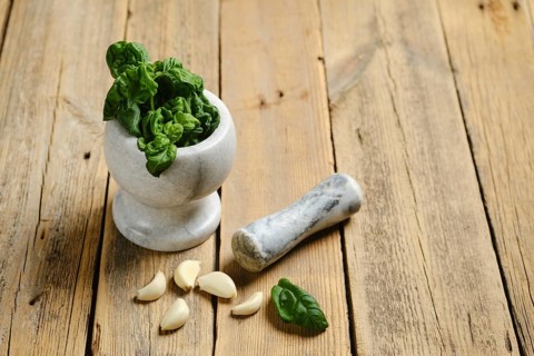 Basil leaves are placed in a mortar with a pestle beside it. Garlic cloves sit on the wooden table.
