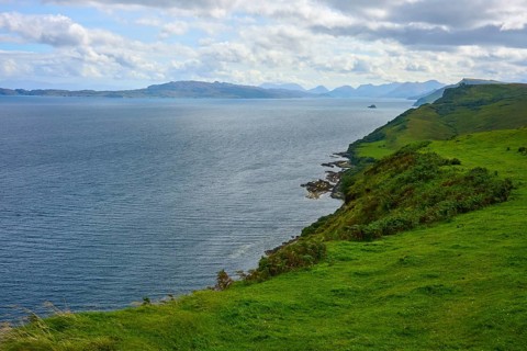 Green coastal landscape with views over sea and distant mountains under cloudy sky, Isle of Skye,