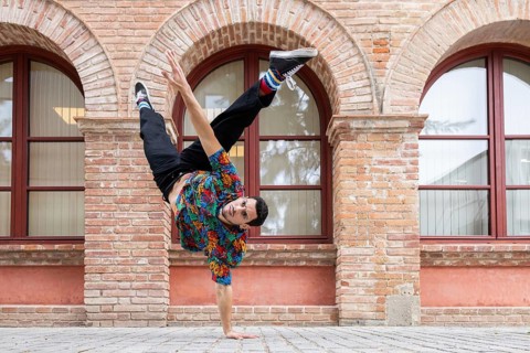 A vibrant Cuban dancer executes a dynamic handstand against a brick wall backdrop. His colorful