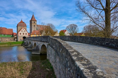 Stone bridge across a river to a village with a church and historic buildings under blue sky,