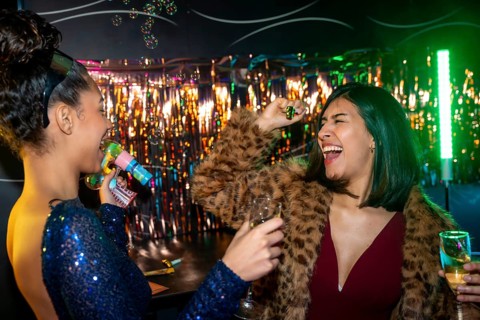 Two diverse young women laugh and dance at a vibrant party, one blowing bubbles with a toy gun